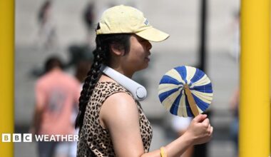 A woman cools herself with a handheld fan whilst walking in the street.