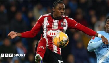 Wigan Athletic player Akeel Higgins, in the red and white of Exeter City last season, tries to control a high ball under challenge from Manchester City player Ryan McAidoo, in sky blue.