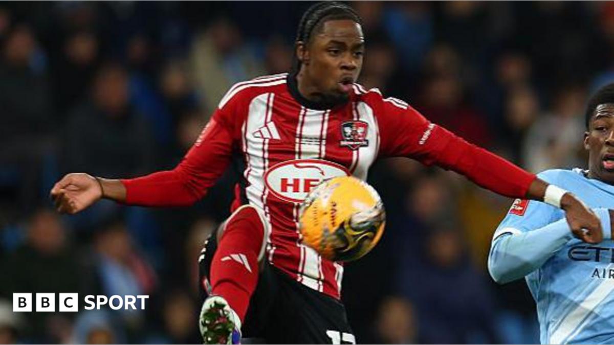 Wigan Athletic player Akeel Higgins, in the red and white of Exeter City last season, tries to control a high ball under challenge from Manchester City player Ryan McAidoo, in sky blue.