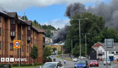 Dense black smoke pours into the sky from an apparent fire behind trees and low-rise buildings beside a curving road. Several cars are driving along the street, while brick apartment blocks line the left side of the image. The smoke contrasts sharply against patches of blue sky and white cloud overhead.