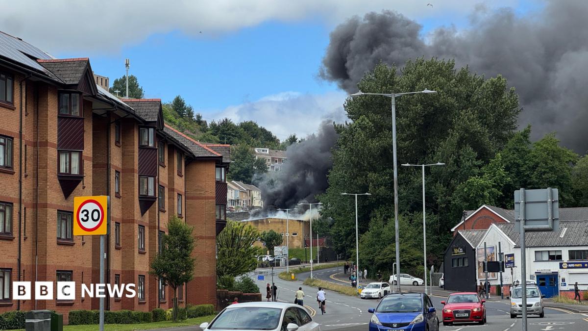 Dense black smoke pours into the sky from an apparent fire behind trees and low-rise buildings beside a curving road. Several cars are driving along the street, while brick apartment blocks line the left side of the image. The smoke contrasts sharply against patches of blue sky and white cloud overhead.
