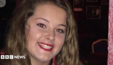 Emily smiles at the camera. She has long wavy brown hair and red lipstick on.