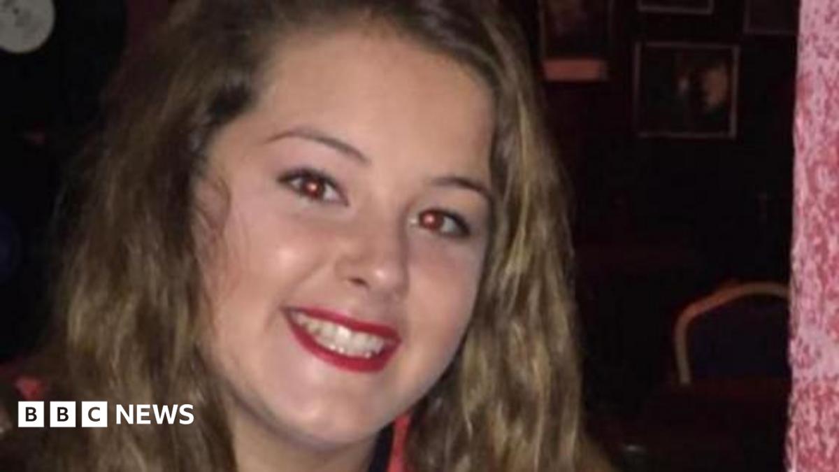 Emily smiles at the camera. She has long wavy brown hair and red lipstick on.