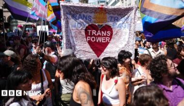 Dozens of people are seen gathered in the background and foreground at the march. A banner is held up by some people in the middle of the photo that reads "Trans joy, trans power, trans rage"