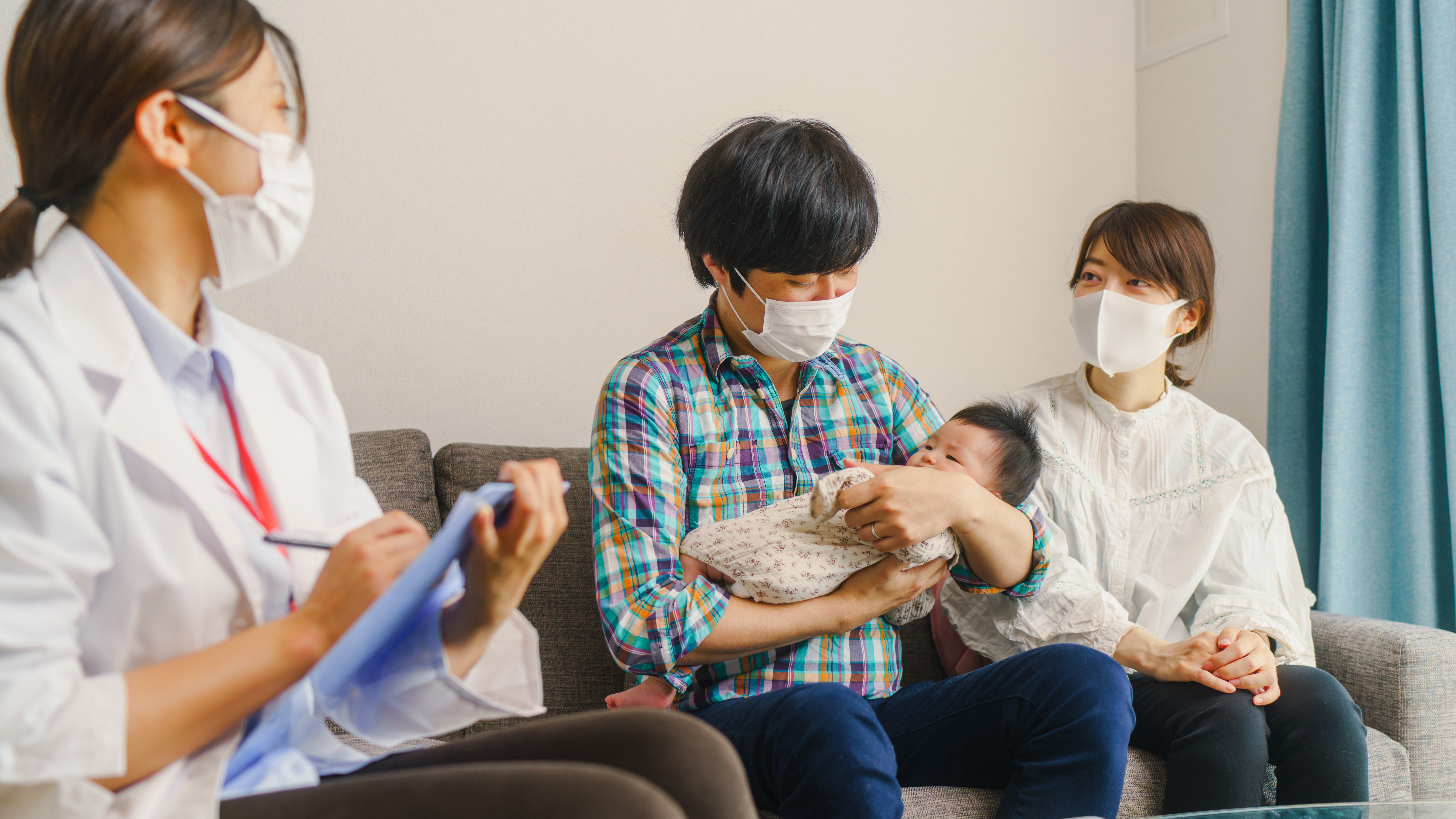 photo of two parents wearing face masks while sitting on a couch across from a masked nurse taking notes. The father holds a swaddled baby.