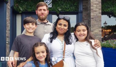 A family photo, a man with brown hair and beard and a woman with dark hair and sunglasses on her head smiling with their three children, all have brown hair.