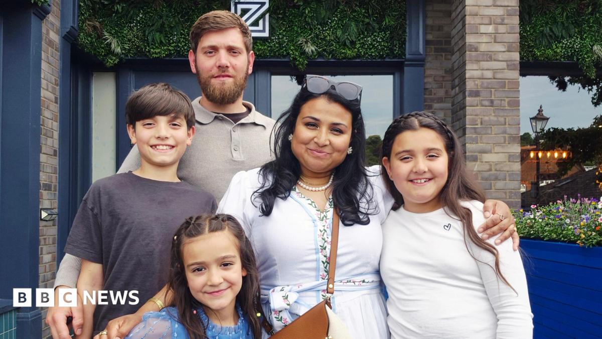 A family photo, a man with brown hair and beard and a woman with dark hair and sunglasses on her head smiling with their three children, all have brown hair.