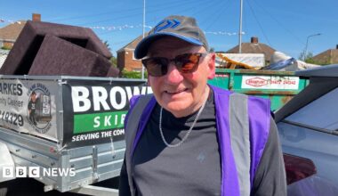 An elderly man wearing a baseball cap, tinted sunglasses and a hi-vis vest over his T-shirt smiles for a photograph.