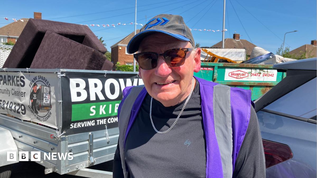 An elderly man wearing a baseball cap, tinted sunglasses and a hi-vis vest over his T-shirt smiles for a photograph.