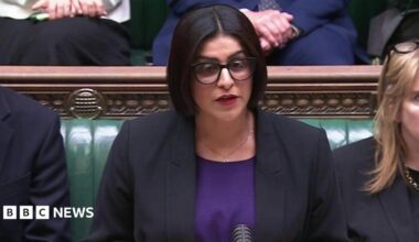 Mahmood wears a black jacket over a royal purple top. She wears her black hair in a chin length bob, with spectacles and red lipstick.