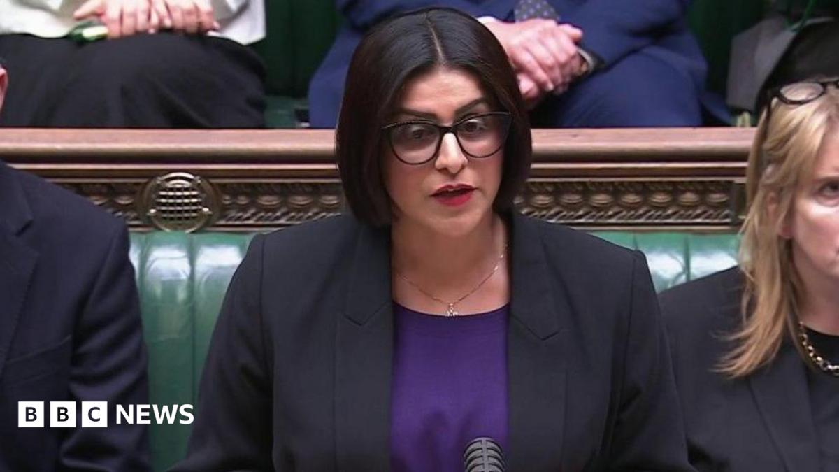 Mahmood wears a black jacket over a royal purple top. She wears her black hair in a chin length bob, with spectacles and red lipstick.