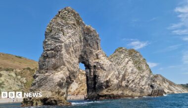 A large rock cliff arch sticks out into the sea. People can be seen on the beach through the arch. There are cliffs behind topped with browning grass. The sky is blue.
