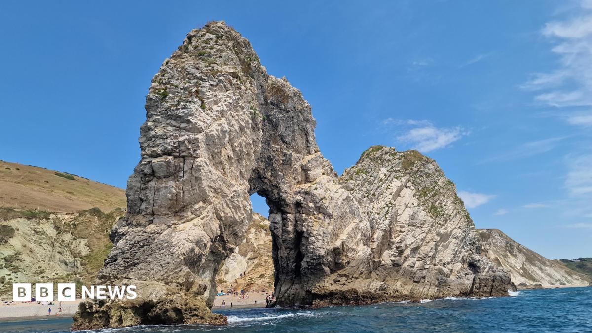 A large rock cliff arch sticks out into the sea. People can be seen on the beach through the arch. There are cliffs behind topped with browning grass. The sky is blue.