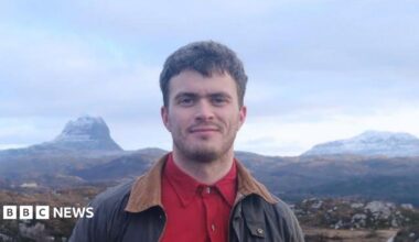 Matthew is wearing a red shirt and brown jacket with a wide collar. Behind him are mountains and sky.