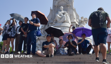 A group of people shield from the sun under umbrellas next to the Victoria Memorial outside Buckingham Palace