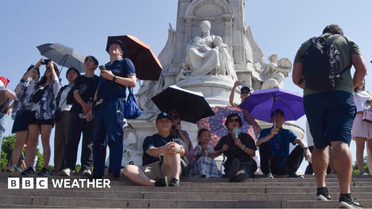 A group of people shield from the sun under umbrellas next to the Victoria Memorial outside Buckingham Palace