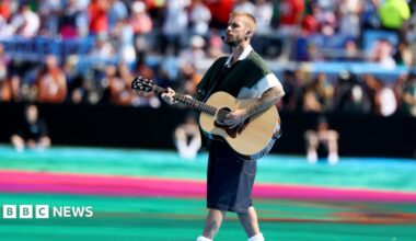 Justin Bieber carrying and playing an acoustic guitar walks across a large stadium field during a live performance. He wears a dark green short-sleeved top layered over a white shirt, knee-length dark shorts, white socks. A headset microphone and in-ear monitors are visible, and tattoos can be seen on both arms and one lower leg. The field is brightly coloured with turquoise and pink sections, while a large, out-of-focus crowd fills the stadium seating in the background. The event is the FIFA World Cup 2026.