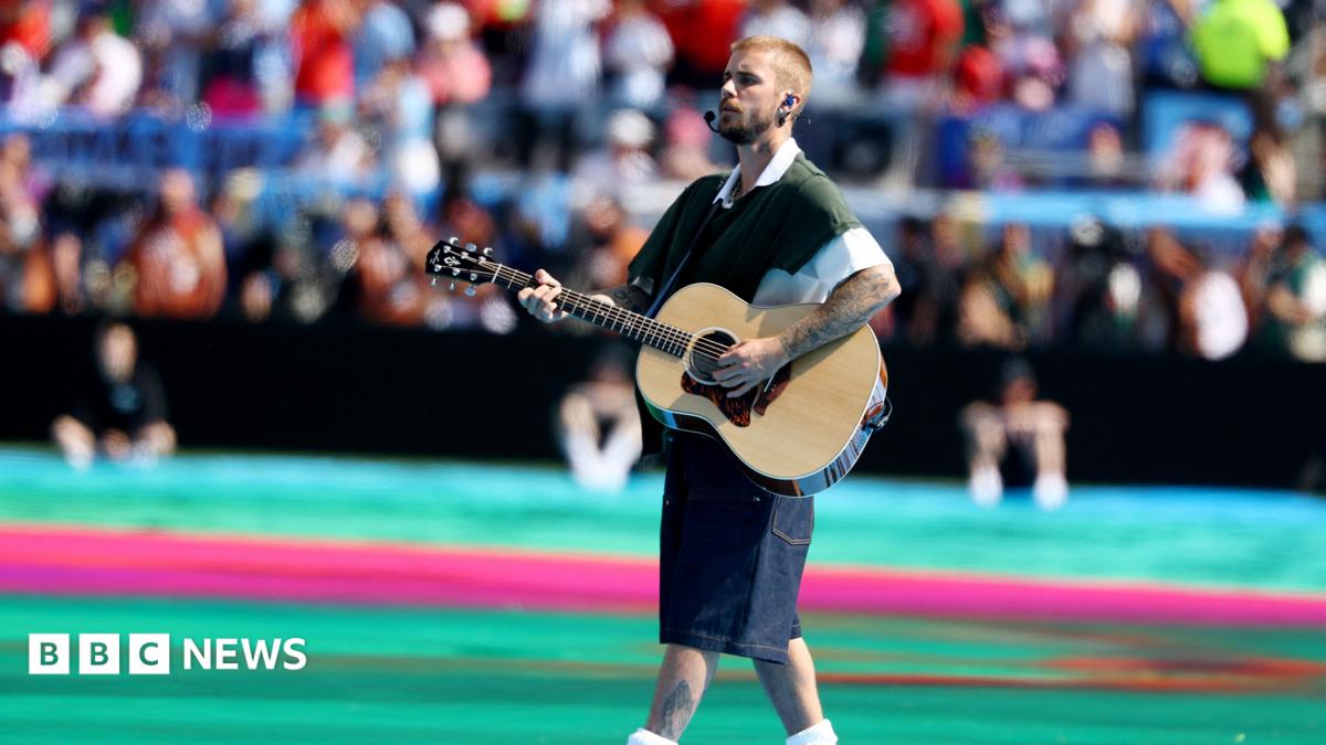 Justin Bieber carrying and playing an acoustic guitar walks across a large stadium field during a live performance. He wears a dark green short-sleeved top layered over a white shirt, knee-length dark shorts, white socks. A headset microphone and in-ear monitors are visible, and tattoos can be seen on both arms and one lower leg. The field is brightly coloured with turquoise and pink sections, while a large, out-of-focus crowd fills the stadium seating in the background. The event is the FIFA World Cup 2026.