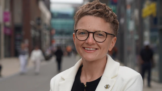 Bev Craig has short red hair, and is wearing glasses and a white jacket over a black blouse