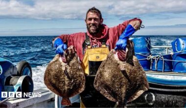 Buck Bennett is standing on a fishing boat holding a plaice in each hand up to show the camera. He is smiling and is wearing rubber gloves and waterproof dungarees over a sweatshirt. The sky is blue with slight cloud and the wake of the boat is white against the dark blue sea.