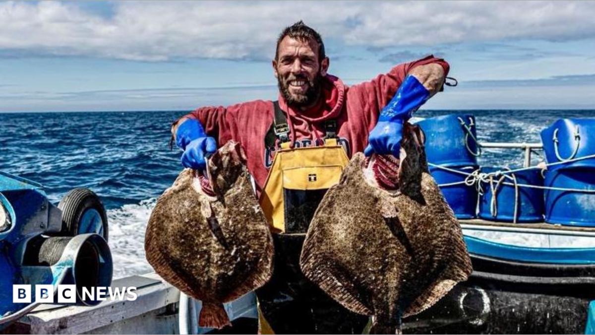 Buck Bennett is standing on a fishing boat holding a plaice in each hand up to show the camera. He is smiling and is wearing rubber gloves and waterproof dungarees over a sweatshirt. The sky is blue with slight cloud and the wake of the boat is white against the dark blue sea.
