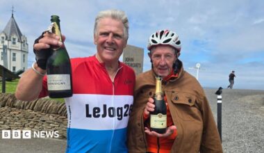 Eddie McGourley and Stephen Foster stand in front of a sign reading 'John O'Groats' with a bottle of champagne each. The sky is cloudy and blue and there is a white-and-grey building behind them. Stephen on the left has grey hair and is wearing a red, white and blue cycling top. Eddie on the right, wears a white cycling helmet and brown jacket buttoned over a red cycling top.