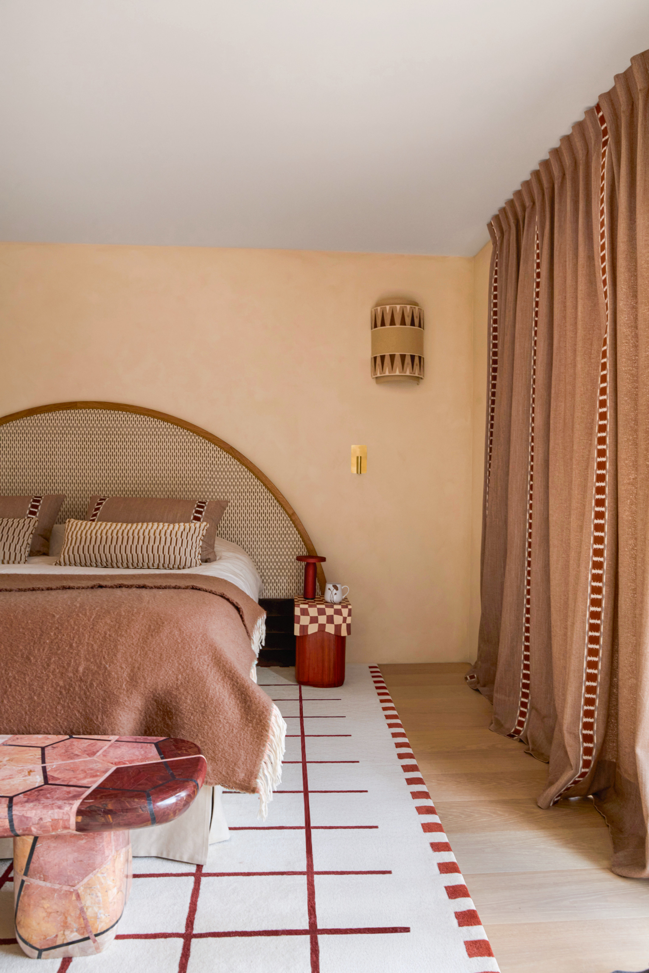 Bedroom with peach textured walls, terracotta curtains, curved oversized headboard, terracotta throw, pink and red stone bench, white and red gridded rug