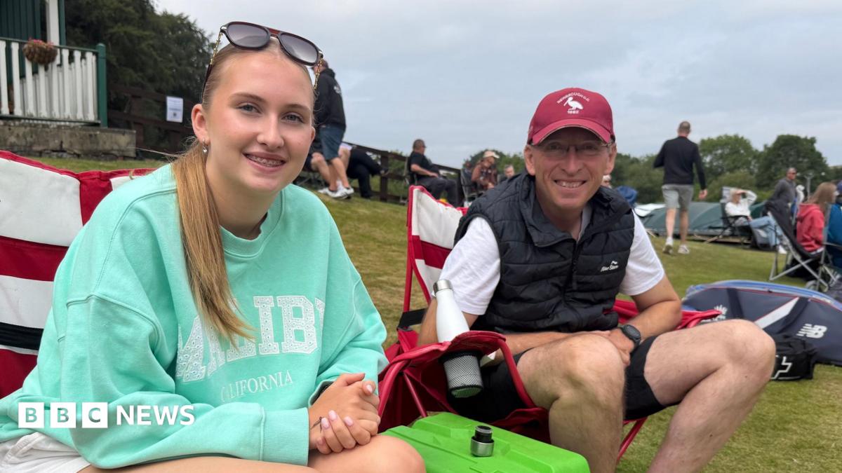 Martin and Poppy sitting on red and white camping chairs and smiling into the camera. Poppy is closest to the camera with light brown hair tied back in a long ponytail. She has sunglasses on top of her head and braces. She is wearing a turquoise jumper with writing on it and has her hands clasped in her lap. Her dad, Martin sits on the right and is hearing a red cap and black bodywarmer on top of a white t-shirt and shorts. There are people sitting around them on the grassy hill.