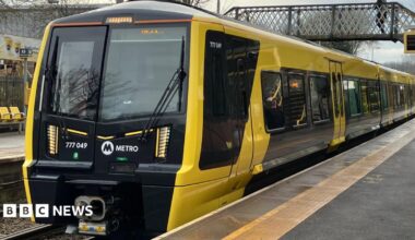 The Merseyrail train is black and yellow, and has stopped at a platform.