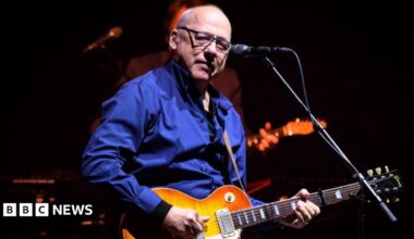 Mark Knopfler performing onstage. He is a bald man with glasses, wearing a blue shirt, and playing a guitar. A microphone is in front of him.