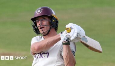 Calvin Harrison of Northamptonshire swings a cricket bat during a County Championship match. He is wearing a white Northamptonshire jersey and a claret helmet with the Northamptonshire rose emblem.