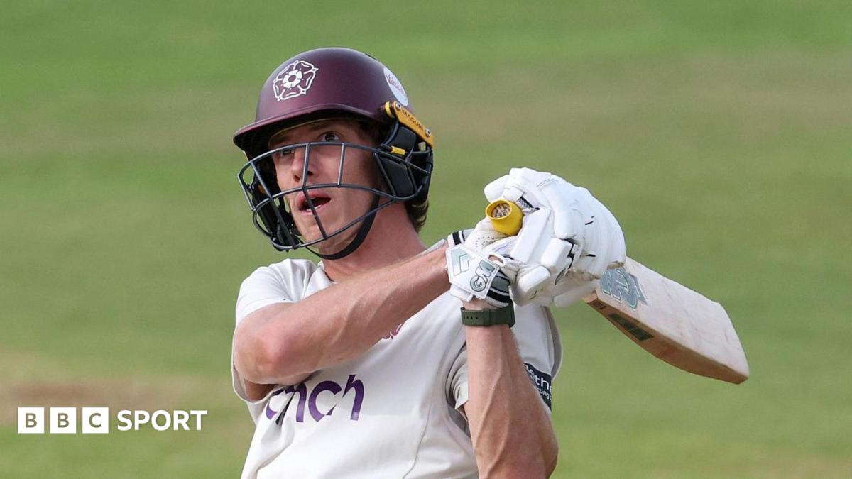 Calvin Harrison of Northamptonshire swings a cricket bat during a County Championship match. He is wearing a white Northamptonshire jersey and a claret helmet with the Northamptonshire rose emblem.
