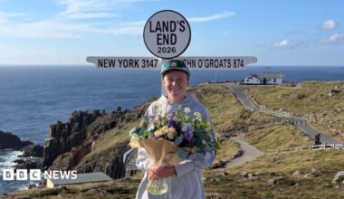 A picture of Peggy Keenor-Crome at the finish line in Land's End. She is wearing a green cycling outfit, a green helmet and is posing with her bike.