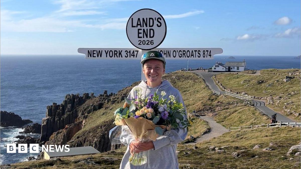A picture of Peggy Keenor-Crome at the finish line in Land's End. She is wearing a green cycling outfit, a green helmet and is posing with her bike.