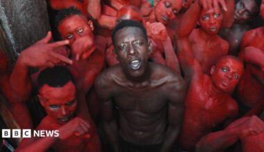 A topless Empereur stands in the centre of a group of young men, who are painted in red.
