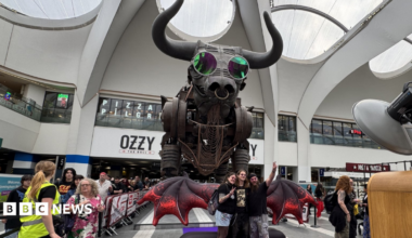 The large mechanical Ozzy the Bull statue is pictured in Birmingham New Street, wearing round pink glasses. An inflatable pair of red wings are positioned in front of him, and a group of p