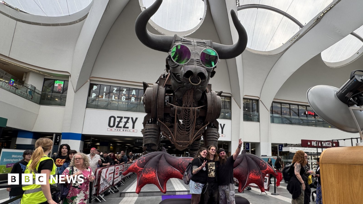 The large mechanical Ozzy the Bull statue is pictured in Birmingham New Street, wearing round pink glasses. An inflatable pair of red wings are positioned in front of him, and a group of p