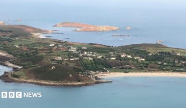 An aerial photo of St Mary's on a sunny day. The island has fields and houses and a long white sand beach can be seen. There are further small islands beyond.