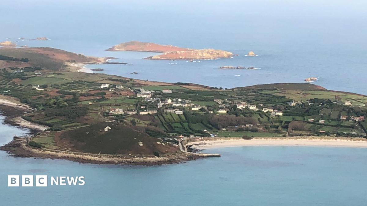 An aerial photo of St Mary's on a sunny day. The island has fields and houses and a long white sand beach can be seen. There are further small islands beyond.