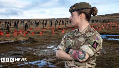 Rhian Brown looks at a display of poppies. Rhian is wearing military fatigues and a beret and has her arms crossed looking away from the camera.