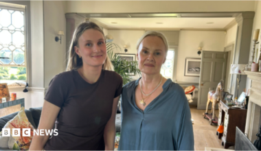 A recent image of Imke, who is now in her 20s, and her mother Caroline, who are looking to camera standing in a large living room with a bay window to the left and a fireplace, desk and rocking horse to the right.