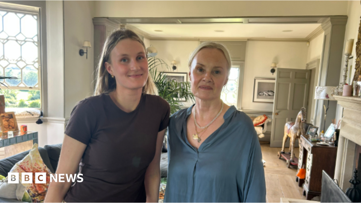 A recent image of Imke, who is now in her 20s, and her mother Caroline, who are looking to camera standing in a large living room with a bay window to the left and a fireplace, desk and rocking horse to the right.