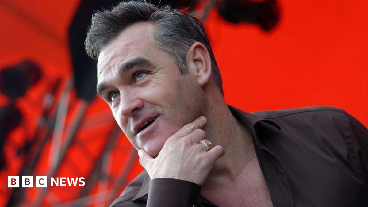 Head and shoulders of Morrissey on stage with his right hand thumb and index finger cupping his chin. He is wearing a gold signet ring, a black shirt and the background is lit red with scafolding