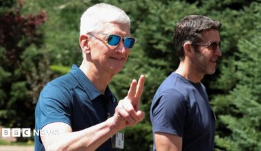 Apple CEO Tim Cook walking outside, throwing up 'peace' fingers, wearing a blue polo shirt and reflective sunglasses. John Ternus, who will replace Cook as CEO later this year, walks beside him, wearing a blue t-shirt and sunglasses.