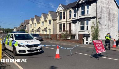 A police car is seen along a road, with police tape and a red "Road Closed" sign