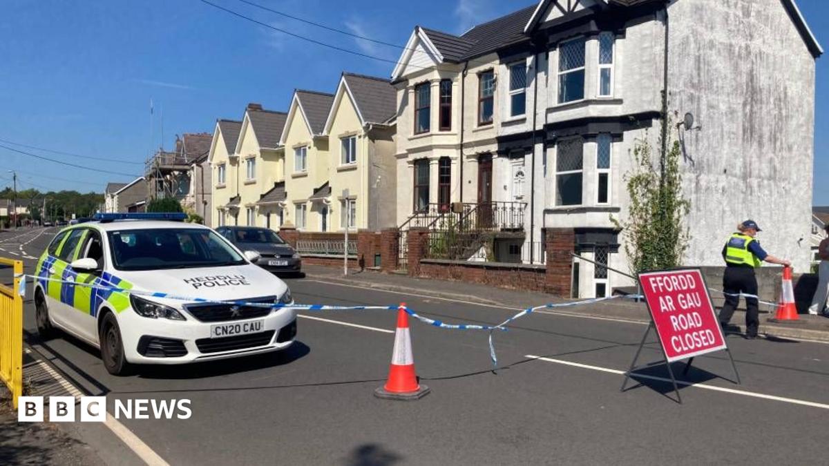 A police car is seen along a road, with police tape and a red "Road Closed" sign