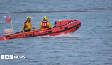 An RNLI boat on the sea at West Mersea. It is orange and has at least two people clad in yellow hi-vis and yellow helmets on board.