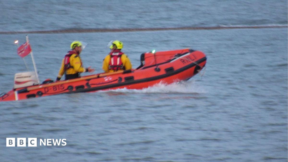An RNLI boat on the sea at West Mersea. It is orange and has at least two people clad in yellow hi-vis and yellow helmets on board.