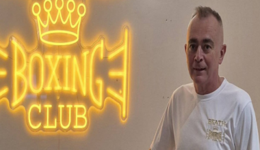Jon-Paul Hynd standing in front of a white wall with the words Beath Boxing Club in large glowing yellow letters. He is wearing a t-shirt with the same logo on it.