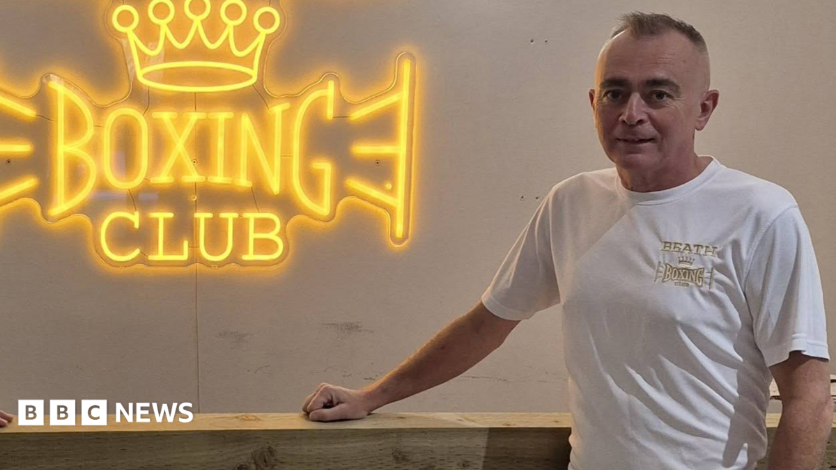 Jon-Paul Hynd standing in front of a white wall with the words Beath Boxing Club in large glowing yellow letters. He is wearing a t-shirt with the same logo on it.
